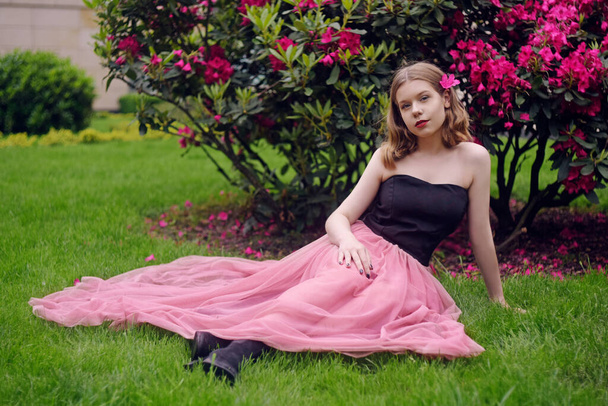 長いピンクのドレスの女の子がピンクの花を背景に芝生の上の庭に座っています。短髪のブロンド、ファッショナブルなプリンセス。彼女の髪の花,カメラを見て,笑顔. - 写真・画像