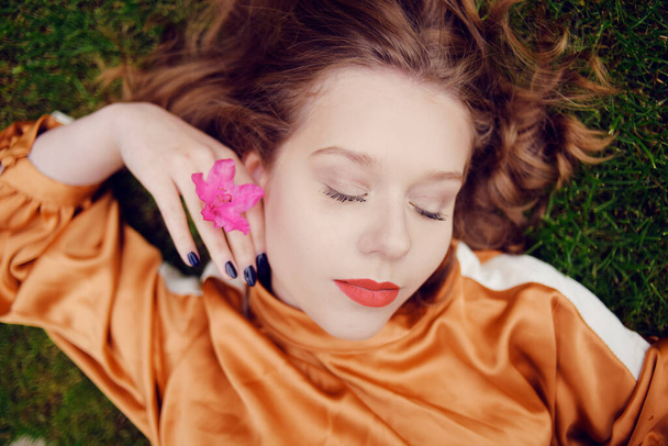 A girl with healthy beautiful hair lies on the grass and looks at the camera. Girl with perfect skin and natural care. Flower in the hands. Layered with eyes closed, sleeping - Photo, Image