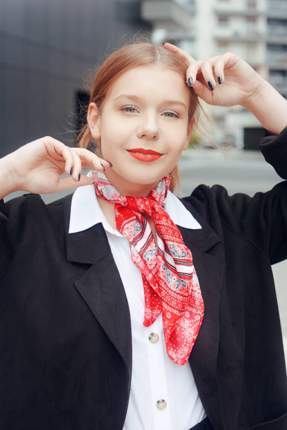 Chica azafata, rubia con cola y labios rojos, en un pañuelo rojo mira a la cámara. Chica con hermoso maquillaje en una camisa blanca y chaqueta posando - Foto, Imagen