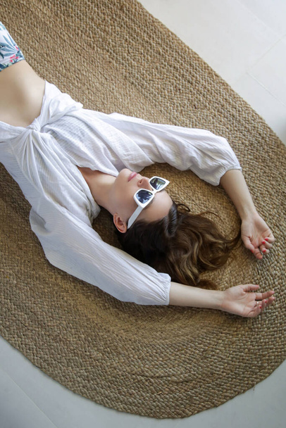 Piękna kobieta leżąca na podłodze. Modna dziewczyna w białej koszuli i białych okularach przeciwsłonecznych. - Zdjęcie, obraz