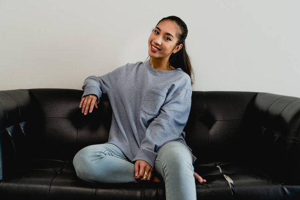 Молода азіатська китаянка виглядає жінкою з світло-блакитним светром, сидячи на чорному дивані і посміхаючись камері
 - Фото, зображення