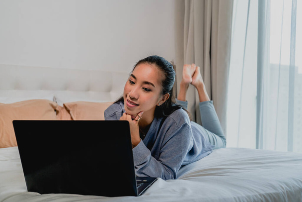 Het Aziatisch chinees uitziende meisje met een lichtblauwe sweater werkt thuis. Liggend op het bed met een laptop. Interieur, huis. - Foto, afbeelding