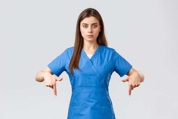 Healthcare workers, prevent virus, covid-19 test screening, medicine concept. Serious professional physician, doctor or nurse in blue scrubs, pointing fingers down, provide coronavirus information - Photo, Image
