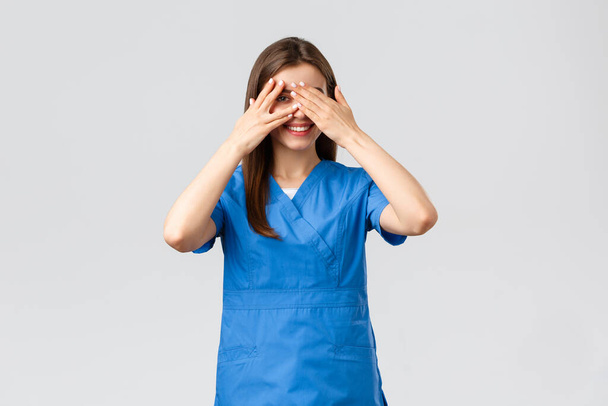 Healthcare workers, prevent virus, insurance and medicine concept. Cute female nurse or doctor in clinic wearing scrubs, cover eyes and peeking through fingers with intrigued smile - Photo, Image