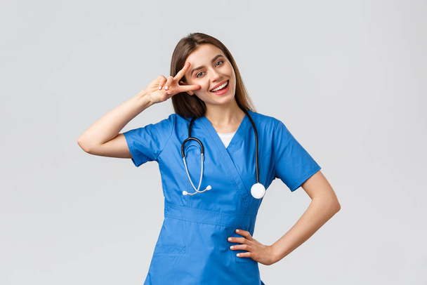 医療従事者、医療、 covid-19と予防接種の概念。青のスクラブで陽気な魅力的な女性看護師や医師,聴診器,平和サインを表示します。,楽観的な滞在,立って灰色の背景 - 写真・画像