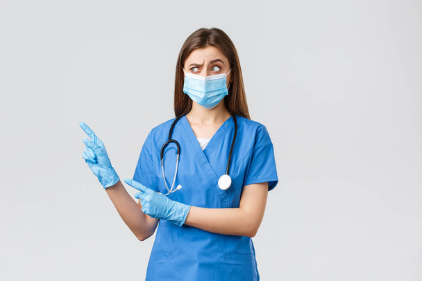 Covid-19 、ウイルス、健康、医療従事者の概念を防止します。青い頭皮で懐疑的な女性看護師,医療用マスクと手袋,疑わしい見て、左上隅を指して - 写真・画像
