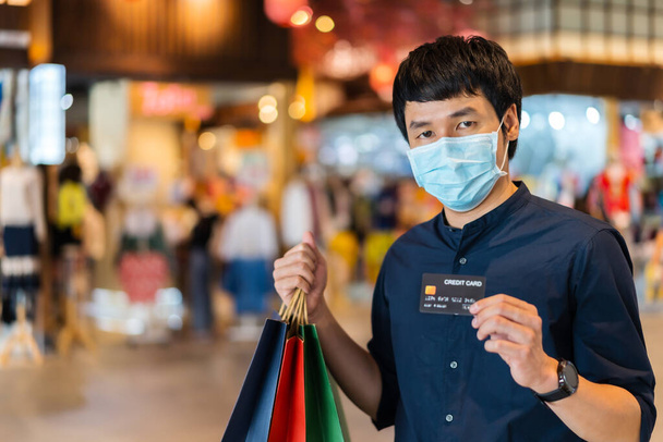 junger asiatischer Mann mit medizinischer Maske und Kreditkarte in einem Einkaufszentrum zur Vorbeugung gegen die Coronavirus-Pandemie (Covid-19). neue normale Konzepte - Foto, Bild