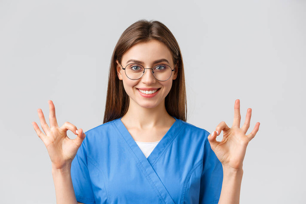 Egészségügyi dolgozók, orvostudomány, biztosítás és a kovid-19 világjárvány koncepciója. Magabiztos és optimista női ápolónő, orvos kék köpeny és szemüveg, mutatnak oké jel, garantálja a legjobb szolgáltatás minőségét - Fotó, kép