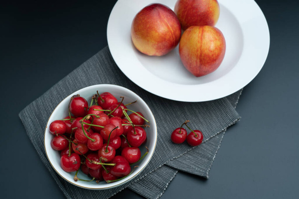 Un tazón de cerezas frescas y un plato de duraznos sobre un fondo negro
 - Foto, Imagen