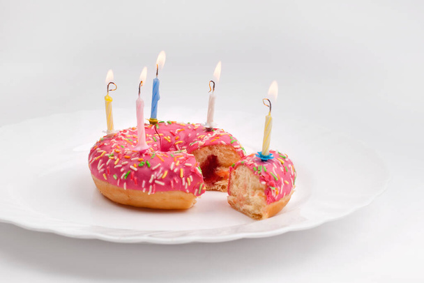 Donut in the form of a birthday cake with burning candles and a cut piece of cake with a candle on a white plate on a white background - Фото, изображение