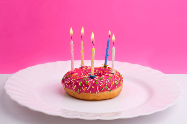Пончик в виде праздничного торта с горящими свечами на белой тарелке на ярко-розовом фоне
 - Фото, изображение