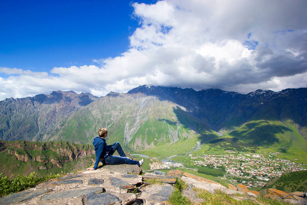 Dziewczyna, kobieta siedzi na kamieniu na szczycie góry, poniżej jest wioska z domami, zielone wysokie góry, wzgórza, głębokie błękitne jasne niebo z ogromnymi białymi chmurami, Kazbek, Stepantsminda, Georgia tło - Zdjęcie, obraz