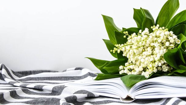 Μπουκέτο με λευκά κρίνα της κοιλάδας στο ανοιχτό βιβλίο για την πετσέτα σε λουρίδα, απαλή εστίαση. Άνοιξη λουλούδια εκπαίδευση, και το διάβασμα, μείνετε στο σπίτι - Φωτογραφία, εικόνα
