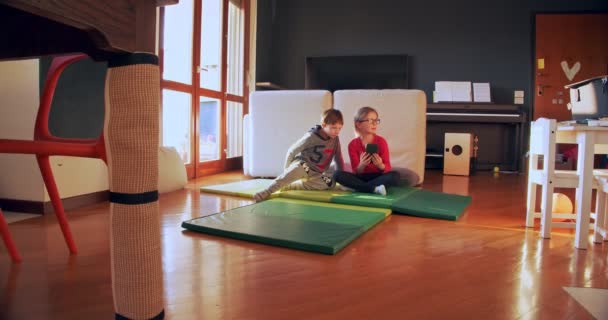 Two children playing together at home with smartphone. Brother and sister watch video on smartphone indoors. Front view medium shot. Kids playing in living room together. Slow motion - Πλάνα, βίντεο