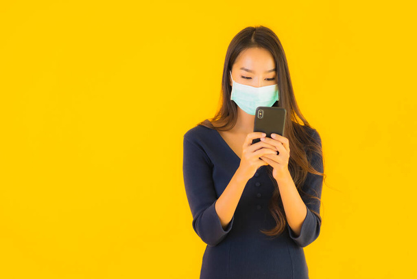 Retrato hermosa joven asiática mujer con máscara y teléfono móvil inteligente o teléfono celular en amarillo aislado fondo
 - Foto, Imagen