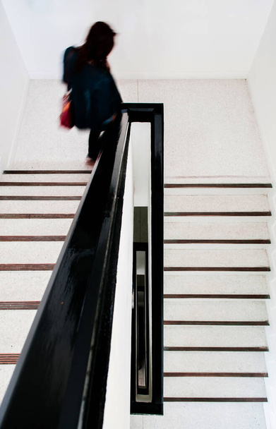 Vrouw loopt naar beneden op eigentijdse stijl wit betonnen trappenhuis met zwart houten leuning schot van bovenaf bekijken. - Foto, afbeelding