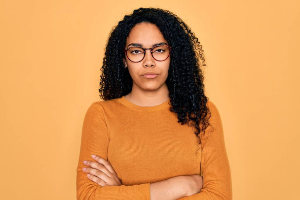 Mladá Afroameričanka v neformálním svetru a brýlích na žlutém pozadí, skeptická a nervózní, nesouhlasící výraz na tváři se zkříženýma rukama. Negativní osoba. - Fotografie, Obrázek