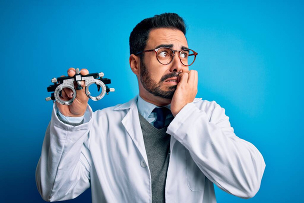 Joven hombre óptico guapo con barba sosteniendo gafas de optometría sobre fondo azul mirando estresado y nervioso con las manos en la boca mordiendo clavos. Problema de ansiedad
. - Foto, imagen