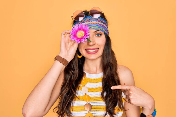 Mavi gözlü genç, güzel hippi kadın güneş gözlüğü takıyor pembe çiçekleri gözünün üstünde tutuyor ve kendini işaret ediyor. - Fotoğraf, Görsel