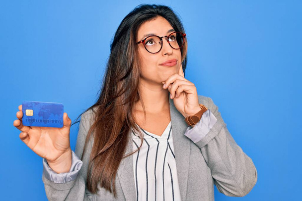 Joven mujer de negocios hispana sosteniendo tarjeta de crédito sobre fondo azul aislado cara seria pensando en la pregunta, idea muy confusa
 - Foto, imagen