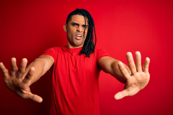 Joven hombre afro afroamericano guapo con rastas usando camiseta casual roja haciendo gesto de stop con palmas de las manos, expresión de enojo y frustración
 - Foto, Imagen