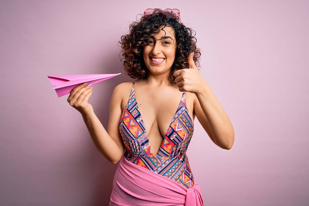 Giovane bella donna araba riccia in vacanza indossando costume da bagno in possesso di carta rosa aeroplano felice con grande sorriso facendo segno ok, pollice in su con le dita, segno eccellente
 - Foto, immagini