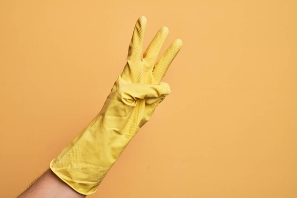 Hand of caucasian young man with cleaning glove over isolated yellow background counting number 3 showing three fingers - Foto, immagini