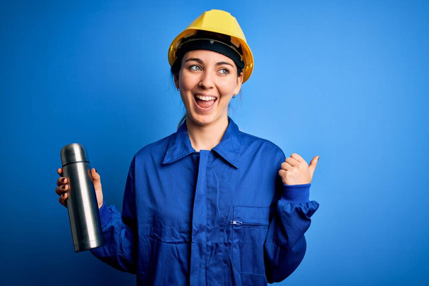 Νεαρή όμορφη μελαχρινή εργάτρια γυναίκα φορώντας κράνος ασφαλείας κρατώντας θερμό με νερό δείχνοντας και δείχνοντας με τον αντίχειρα μέχρι το πλάι με χαρούμενο πρόσωπο χαμογελώντας - Φωτογραφία, εικόνα