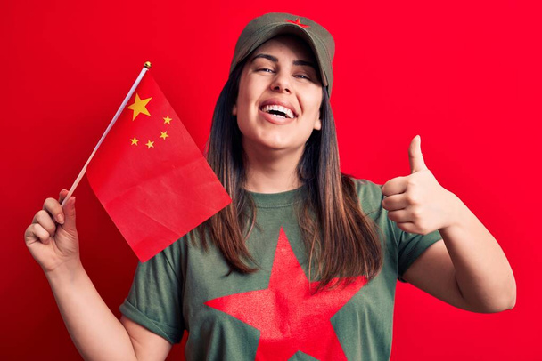 Gyönyörű hazafias nő, piros csillagos kommunista szimbólumos pólóban, porcelánzászlóval a kezében, boldog és pozitív mosollyal, hüvelykujjával felfelé, kiváló munkát végez és jóváhagyási jel. - Fotó, kép