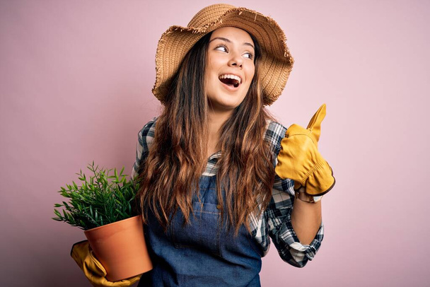 Mladá krásná brunetka farmářka žena nosí zástěru a klobouk drží hrnec s rostlinami ukazuje palcem nahoru na stranu se šťastnou tváří usmívá - Fotografie, Obrázek