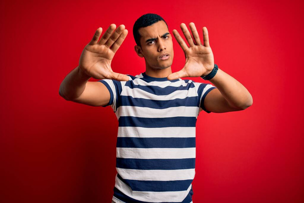 Hombre afroamericano guapo usando casual rayas camiseta de pie sobre fondo rojo haciendo marco usando las manos palmas y los dedos, perspectiva de la cámara
 - Foto, Imagen