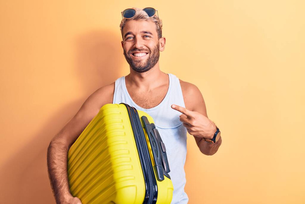 Молодой красивый блондин-турист в отпуске в солнечных очках, держа сумку из самолета, улыбаясь счастливо указывая рукой и пальцем.
 - Фото, изображение