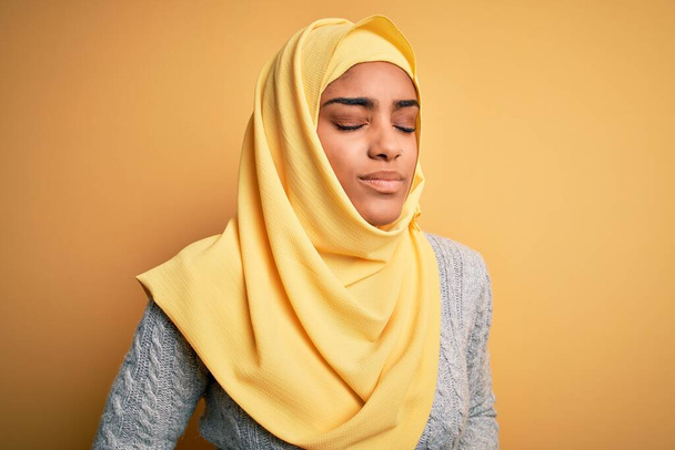 Young beautiful african american girl wearing muslim hijab over isolated yellow background with hand on stomach because indigestion, painful illness feeling unwell. Ache concept. - Fotografie, Obrázek