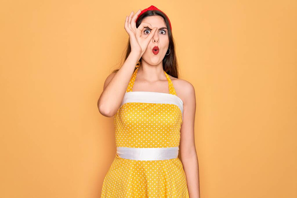 Fiatal szép pin up nő visel 50-es évek divat vintage ruha sárga háttér csinál oké gesztus döbbent arc, szem néz keresztül ujjait. Hitetlen kifejezés.. - Fotó, kép
