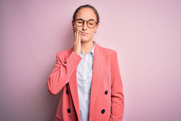 Jeune belle femme d'affaires portant une veste et des lunettes sur fond rose isolé touchant la bouche avec une expression douloureuse à cause de maux de dents ou de maladies dentaires sur les dents. Dentiste - Photo, image