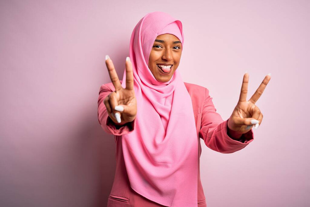 Jonge Afro-Amerikaanse afro-vrouw draagt moslim hijab over geïsoleerde roze achtergrond glimlachend met de tong uit te laten zien vingers van beide handen doen overwinning teken. Nummer twee. - Foto, afbeelding