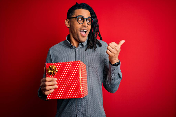 Афроамериканец с дредами, держащий настоящую коробку на красном изолированном фоне, указывая пальцем вверх в сторону, со счастливым лицом, улыбающимся
 - Фото, изображение