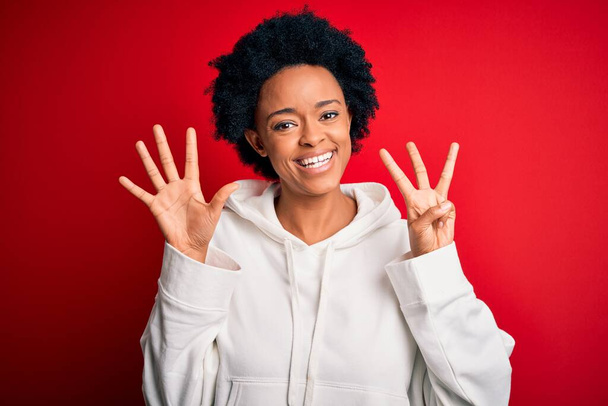 Junge schöne afroamerikanische Afro-Sportlerin mit lockigem Haar, die Sportbekleidung trägt und mit Finger Nummer acht nach oben zeigt, während sie selbstbewusst und glücklich lächelt. - Foto, Bild