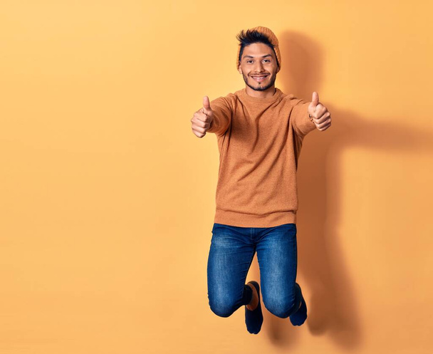 Young handsome latin man wearing casual clothes and wool cap smiling happy. Jumping with smile on face doing ok sign with thumbs up over isolated yellow background - Foto, Bild