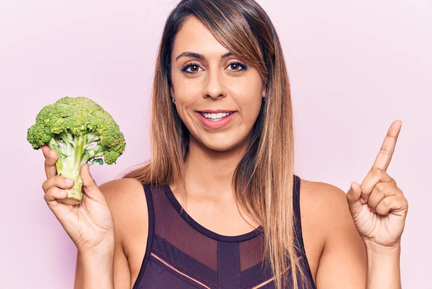 Young beautiful woman holding broccoli smiling happy pointing with hand and finger to the side  - Фото, изображение