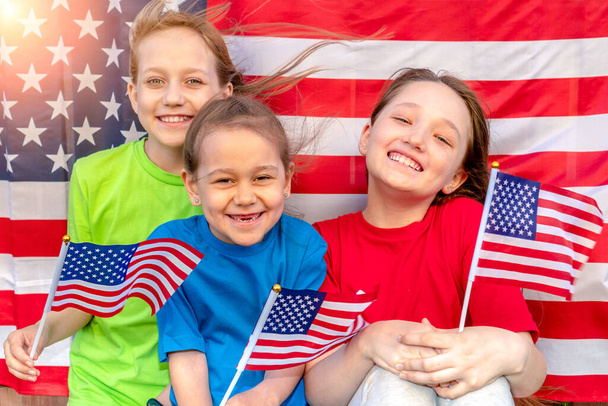 Tři šťastné dívky s americkými vlajkami v rukou se dívají do kamery a usmívají se. Vítr rozvíjí vlasy a vlajky. Den nezávislosti. Vlastenecká dovolená. - Fotografie, Obrázek