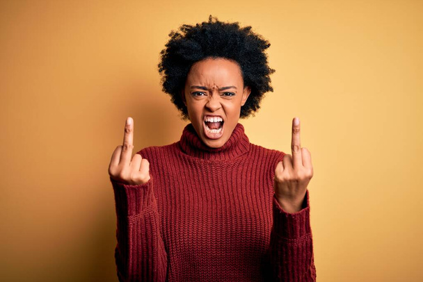Young beautiful African American afro woman with curly hair wearing casual turtleneck sweater Showing middle finger doing fuck you bad expression, provocation and rude attitude. Screaming excited - Photo, Image
