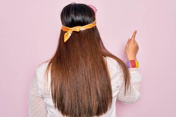 Jeune femme hippie hispanique portant un style boho de la mode et des lunettes de soleil sur fond rose Posant vers l'arrière pointant vers l'avant avec la main du doigt - Photo, image