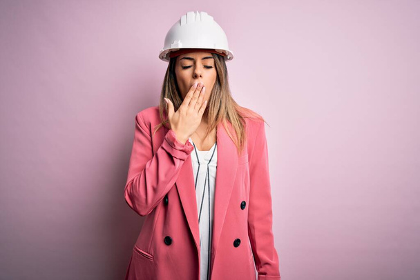 Молодая красивая брюнетка архитектор женщина в защитном шлеме на розовом фоне скучно зевая усталый покрывающий рот рукой. Беспокойство и сонливость
. - Фото, изображение