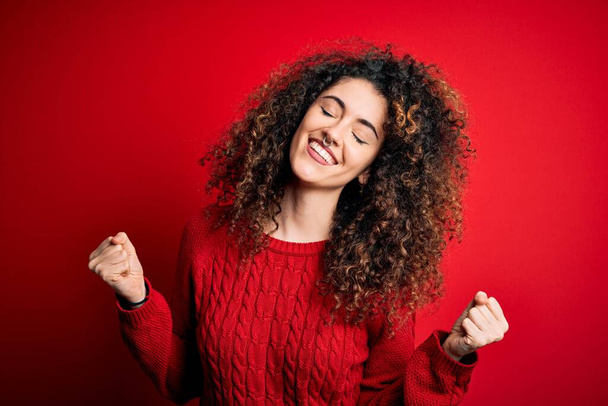 Young beautiful woman with curly hair and piercing wearing casual red sweater very happy and excited doing winner gesture with arms raised, smiling and screaming for success. Celebration concept. - Foto, imagen