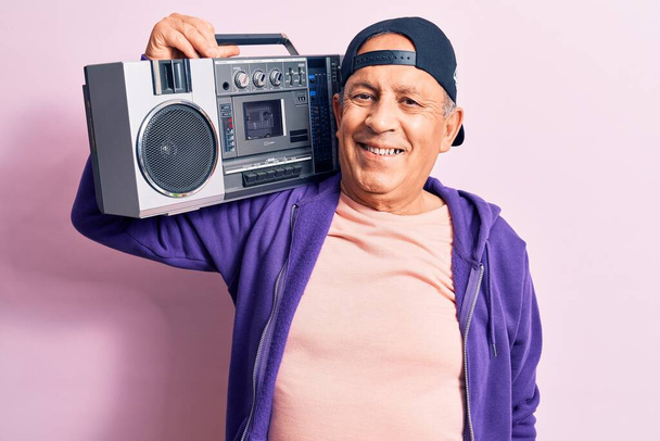 Senior guapo hombre moderno de pelo gris escuchando música usando boombox vintage mirando positiva y feliz de pie y sonriendo con una sonrisa confiada mostrando los dientes
 - Foto, Imagen
