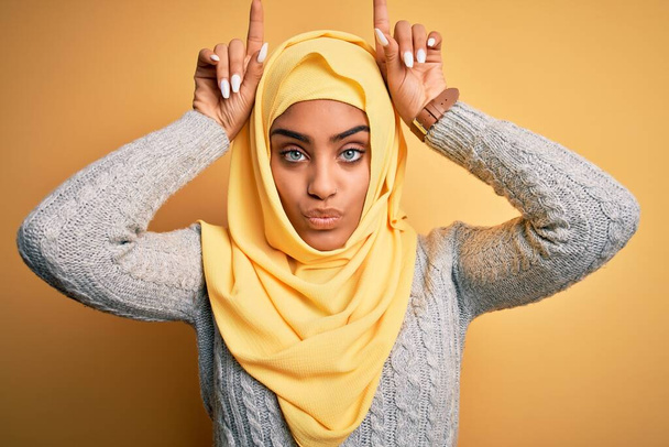 Молодая красивая африканская американская девушка в мусульманском хиджабе на изолированном желтом фоне делает смешной жест с пальцем над головой, как рога быка
 - Фото, изображение