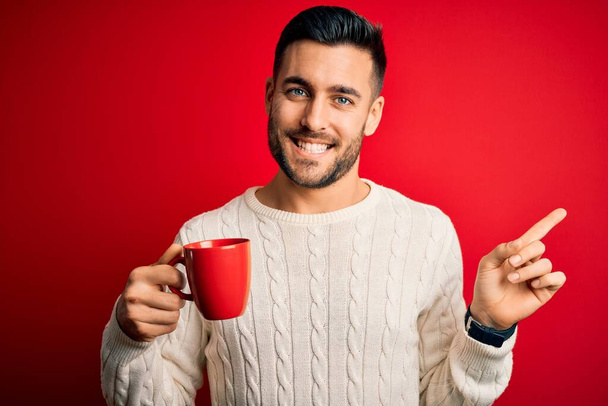 Joven hombre guapo bebiendo una taza de café caliente sobre fondo rojo aislado muy feliz señalando con la mano y el dedo a un lado
 - Foto, imagen