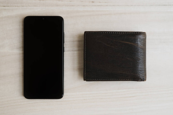 Fekete mobiltelefon és barna bőrtárca a fa asztalon, felülnézetből. Mobil banki koncepció. Online pénztárca   - Fotó, kép