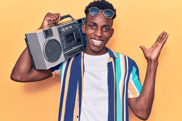 Genç Afrikalı Amerikalı adam elinde teyp ile zaferi kutlayan müzikler dinlerken mutlu bir gülümseme ve ellerini kaldırarak kazanan bir ifade takınıyor.  - Fotoğraf, Görsel